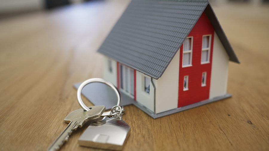 Fotografia de uma chave e chaveiro metalizado junto a uma miniatura de residência de dois andares com paredes nas cores vermelha e branca e telhado cinza, em uma mesa de madeira. Foto: Tierra Mallorca / Unsplash.