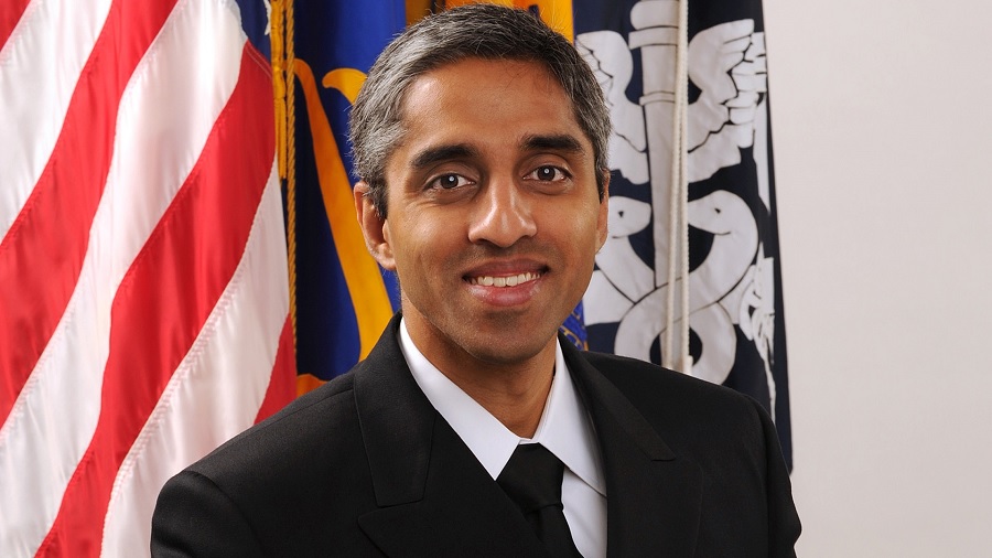 Fotografia, em primeiro plano, de Vivek Murthy sorridente, à frente das bandeiras dos EUA e do Departamento de Saúde e Serviços Humanos americano. Foto: Wikimedia Commons.