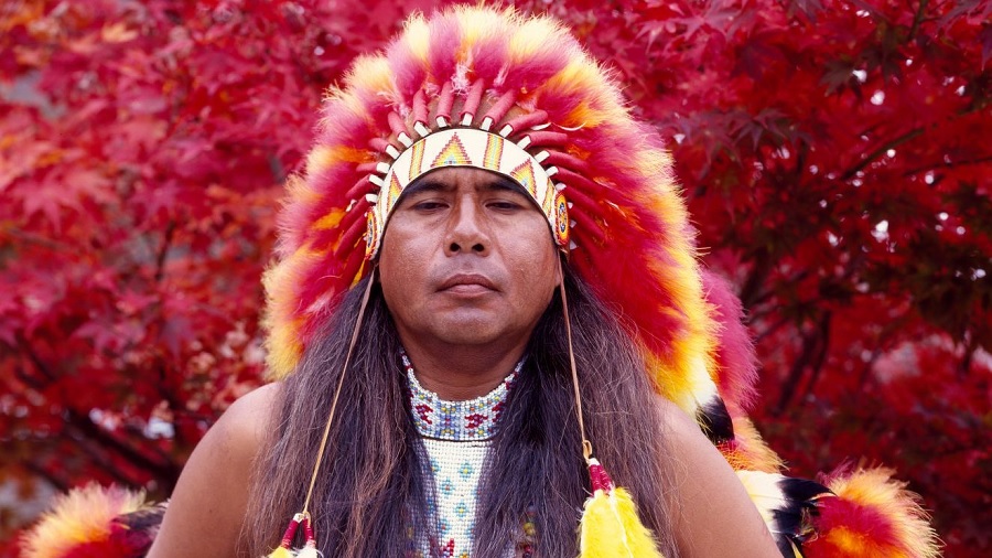 Fotografia de um índio Cherokee usando cocar vermelho e amarelo e cabelos compridos à frente dos ombros e a folhagem vermelha de uma árvore de bordo fechando a composição ao fundo. Imagem: Carol M. Highsmith / Biblioteca do Congresso dos EUA.