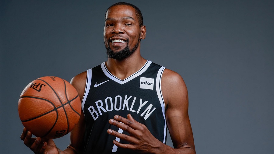 Fotografia, em primeiro plano, de Kevin Durant sorridente e usando uniforme preto e branco do Brooklyn Nets, enquanto segura a bola em uma das mãos, à frente de um fundo cinza. Imagem: Nicole Sweet | USA Today.