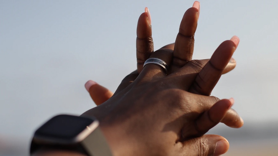 Fotografia mostra duas mãos com os dedos entrelaçados, onde uma aliança é vista no dedo anelar da mão que está com o dorso voltado para a câmera, e um fundo formado por tons de cinza embaçado. Imagem: Goke Obasa / Unsplash.