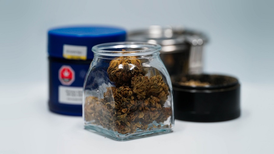 Fotografia mostra um vidro de base quadrada e boca redonda cheio de cannabis e três potes, um azul, um metálico e outro preto, que aparecem no segundo plano, fora do foco, em uma superfície branca e lisa. Photo by 2H Media on Unsplash.