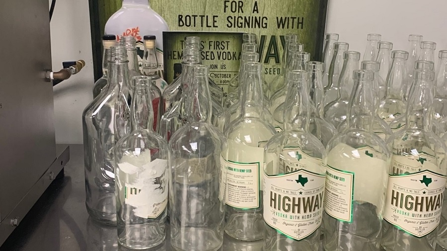Fotografia mostra várias garrafas da vodca Highway Vodka vazias enfileiradas à frente de um banner da destilaria, em uma bancada de inox. Foto: Erika Kwee / Houston Press.