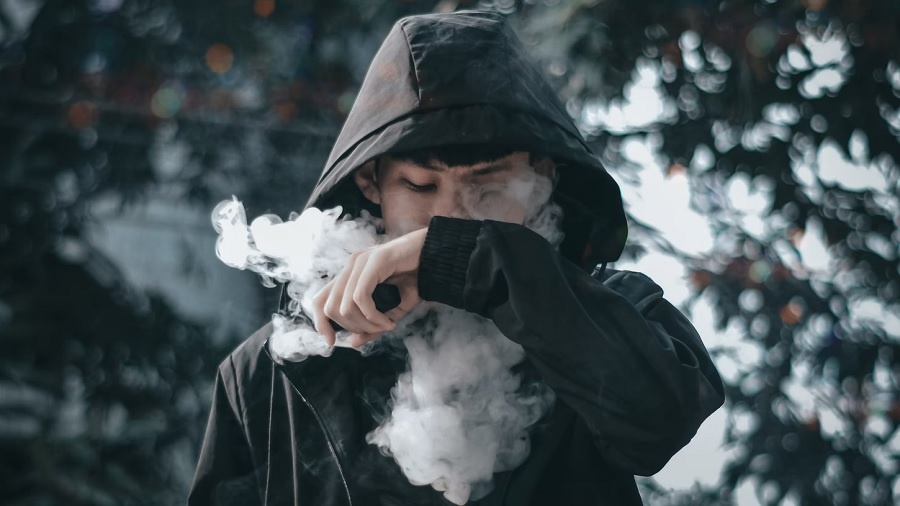 Foto mostra uma pessoa usando casaco com capuz preto e segurando um dispositivo de vaporização da mesma cor, enquanto posiciona o punho à frente da boca e expele uma nuvem de vapor, com o olhar voltado para baixo, e um fundo de árvores e luzes bokeh embaçado. Imagem: Toan Nguyen | Unsplash.