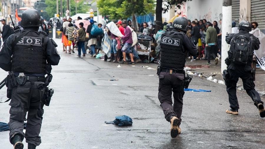Fotografia mostra três policias com uniforme do GOE de costas, enquanto caminham em direção a várias pessoas que estão na região da Cracolândia, estando dois deles empunhando fuzis. Imagem: Daniel Arroyo / Ponte.