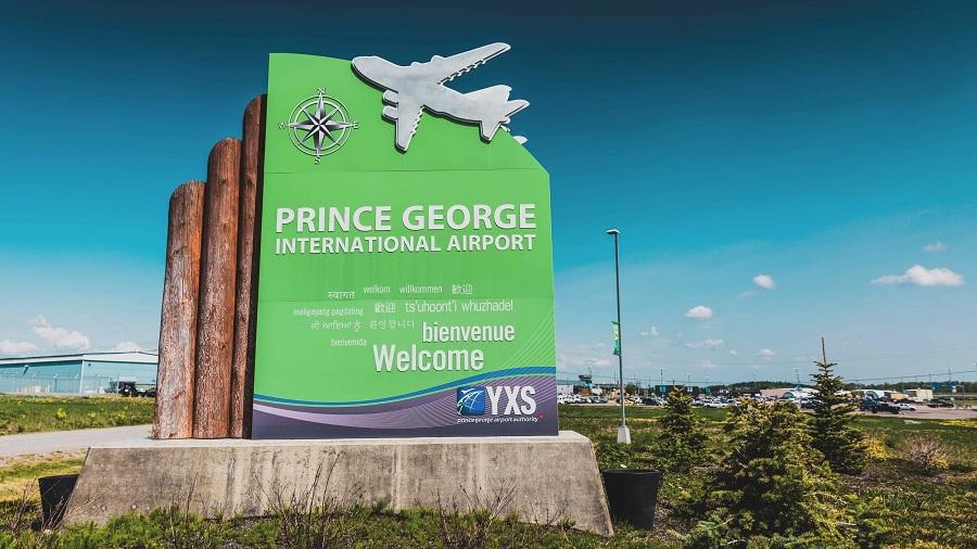Fotografia mostra a área externa do aeroporto de Prince George, onde se vê um banner de “bem-vindo”, com as cores verde e branco predominando e os desenhos de uma rosa dos ventos e um avião, entre vegetação. Foto: Prince George Airport / Divulgação.