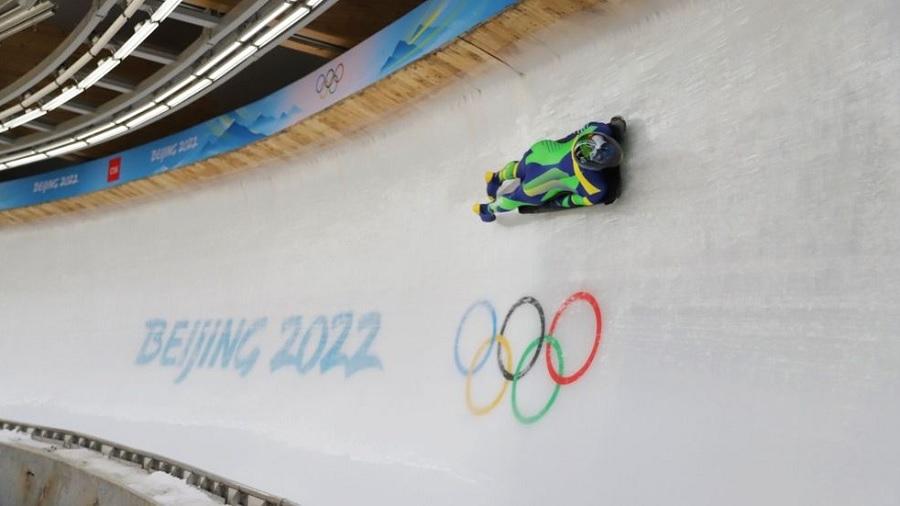 Fotografia mostra a atleta Nicole Silveira descendo a pista de gelo, onde se vê o logo dos Jogos de Pequim 2022 e os aros olímpicos, na modalidade skeleton. Imagem: Alexandre Castelo Branco / COB.