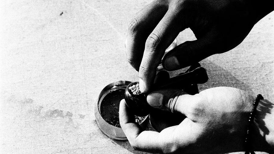 Fotografia, em P&B, mostra as mãos de uma pessoa que segura um pipe de cor escura enquanto ajeita a erva no fornilho com a ponta do indicador, sobre um fundo irregular de cor clara. Foto: Unsplash / Chris Carzoli.