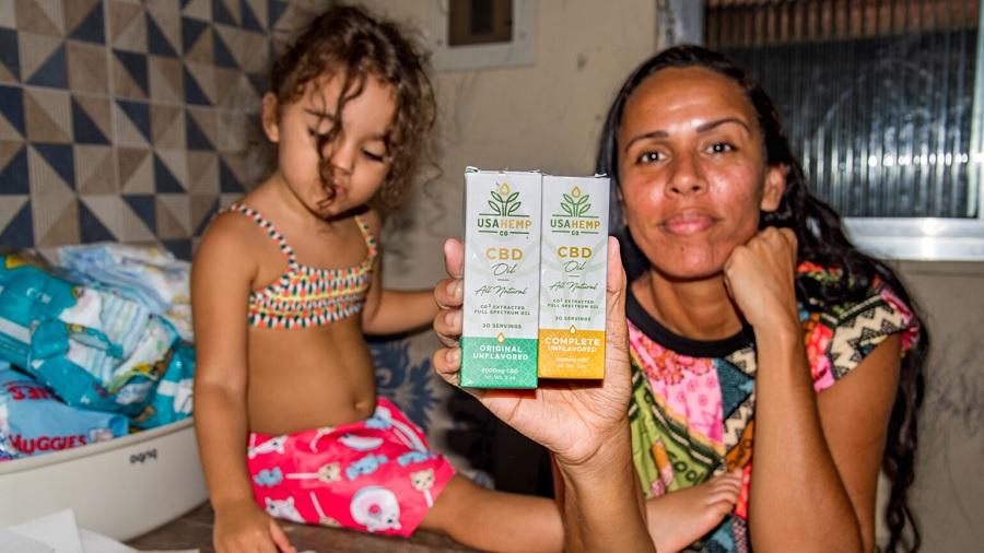 Fotografia mostra Rafaela França segurando duas caixas do óleo de cannabis, uma de cada versão (verde e amarela), próximo à filha Maria, em um ambiente interno. Foto: Selma Souza / Voz das Comunidades.