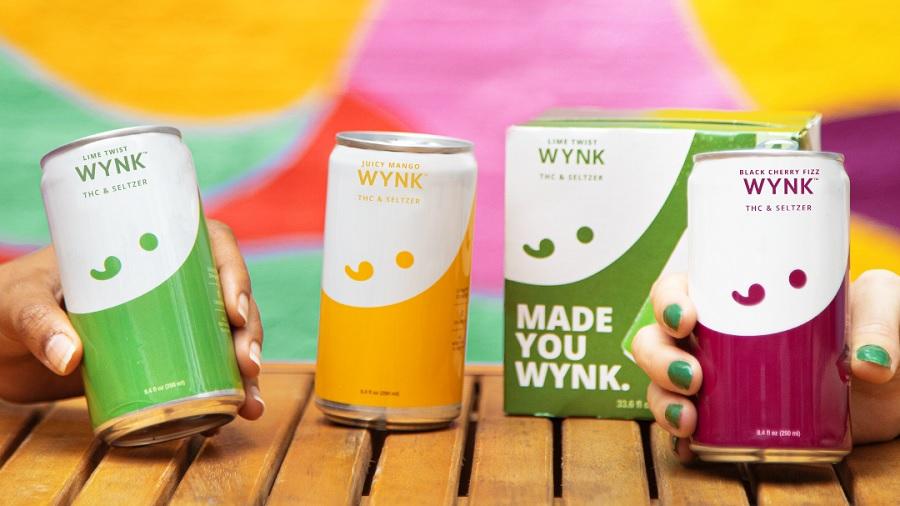 Fotografia mostra uma lata de cada sabor da seltzer Wynk, com duas sendo seguradas por mãos que vêm das laterais da imagem, além de uma caixa da bebida, sobre um deque, e um fundo colorido. Foto: Divulgação.
