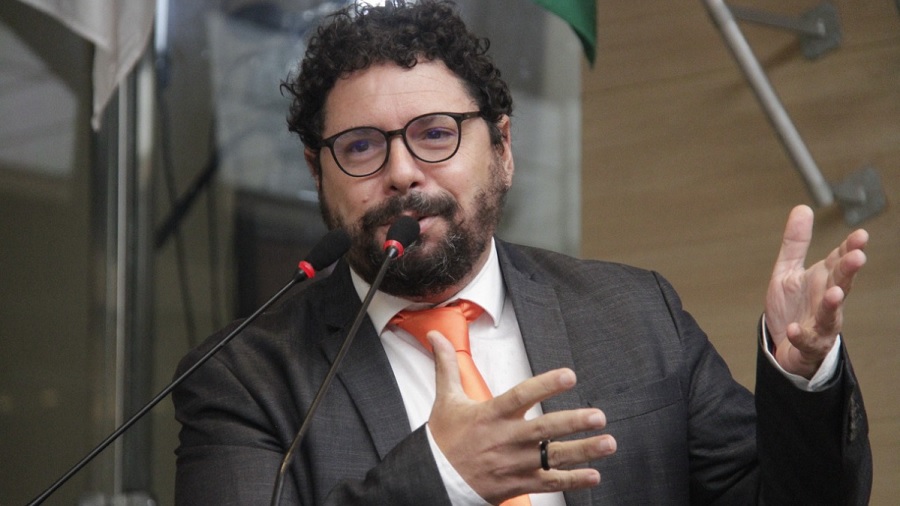 Fotografia em primeiro plano de Ivan Moraes, onde o vereador aparece de terno cinza e gravata salmão e gesticulando enquanto fala aos microfones da tribuna. Imagem: Câmara do Recife.