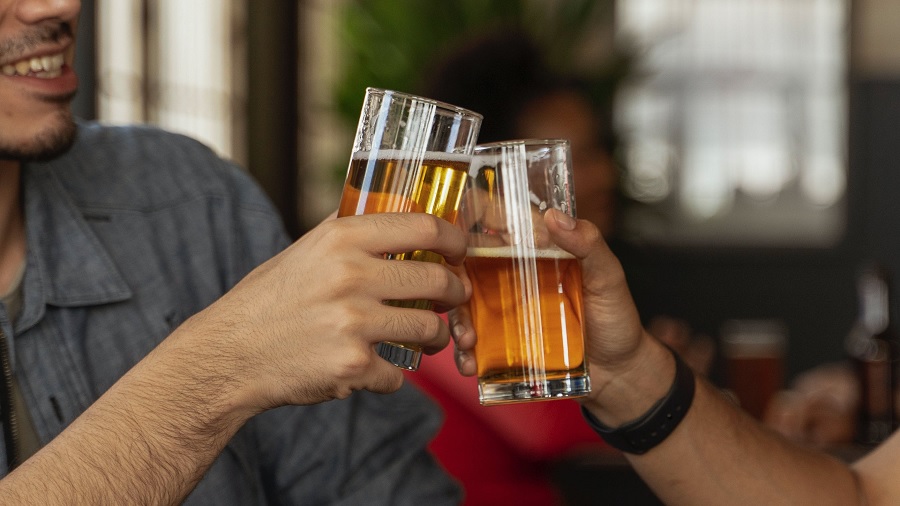 Fotografia mostra as mãos e braços de duas pessoas que batem os copos de cerveja, e parte do corpo de uma delas, que aparece sorridente — ao fundo, fora de foco, se vê outra pessoa sentada a uma mesa. Imagem: Ketut Subiyanto / Pexels.