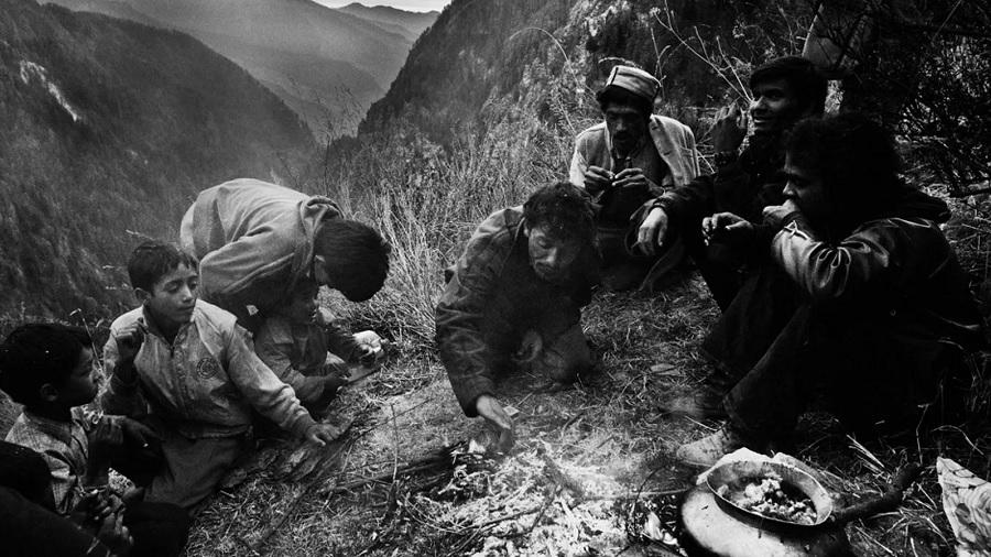 Fotografia, em P&B, mostra um grupo de pessoas reunidas em torno de uma fogueira na encosta de uma colina, em Malana. Imagem: Hari Karagadda.