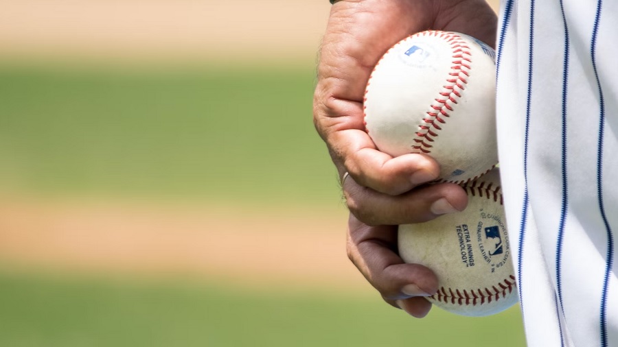 Foto mostra a mão e parte do uniforme, branco e azul, de um jogador de beisebol, que segura duas bolas, e o gramado do campo, ao fundo, fora do foco. Imagem: Jose Morales / Unsplash.