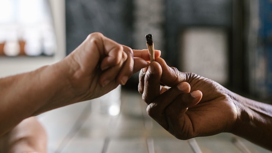 Foto mostra duas mãos vindas das laterais da imagem e se encontrando ao centro, enquanto passam um cigarro de cannabis, em fundo embaçado. Fotografia: Pexels / Rodnae Productions.