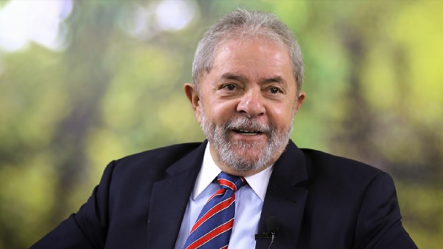 Fotografia do peito para cima de Lula, usando paletó azul-escuro e gravata listrada vermelha e azul, com fundo embaçado em tons de verde. Imagem: Ricardo Stuckert / divulgação.