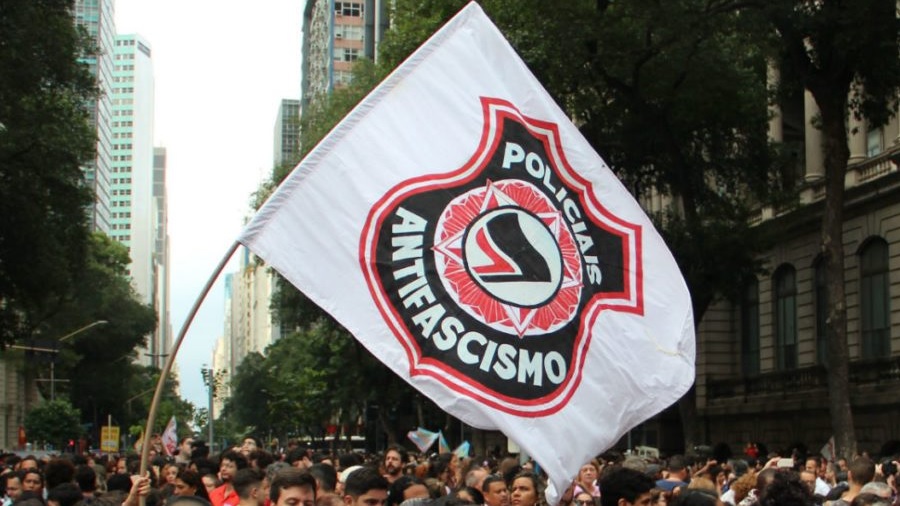 Foto mostra uma bandeira branca com o brasão, em preto e vermelho, do Policiais Antifascismo sendo levantada em meio a uma multidão, e edifícios e árvores, ao fundo.