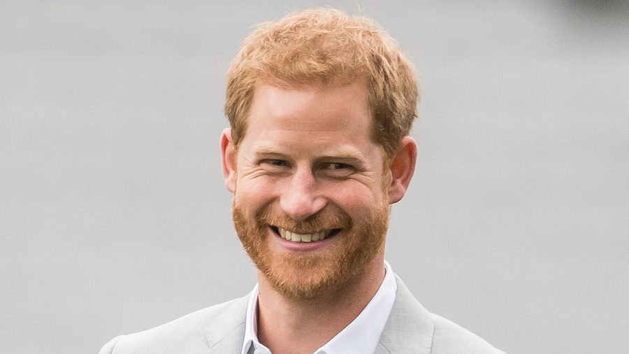 Fotografia mostra o príncipe Harry, dos ombros para cima, sorridente e usando um paletó cinza-claro, e um fundo cinza.
