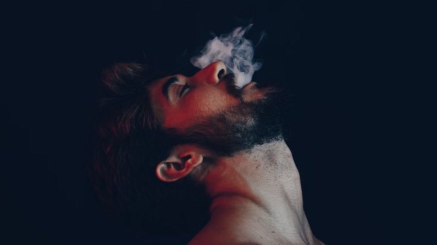 Fotografia mostra um homem, em perfil, dos ombros para cima e com a cabeça voltada para trás, expelindo fumaça e um fundo escuro. Imagem: Unsplash | César Couto.