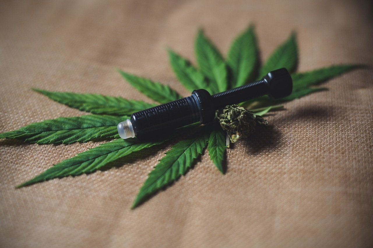 Foto mostra uma seringa preta e um bud de cannabis sobre folhas de cannabis dispostas sobre um tecido bege. Imagem: Pixabay | Julia Teichmann.