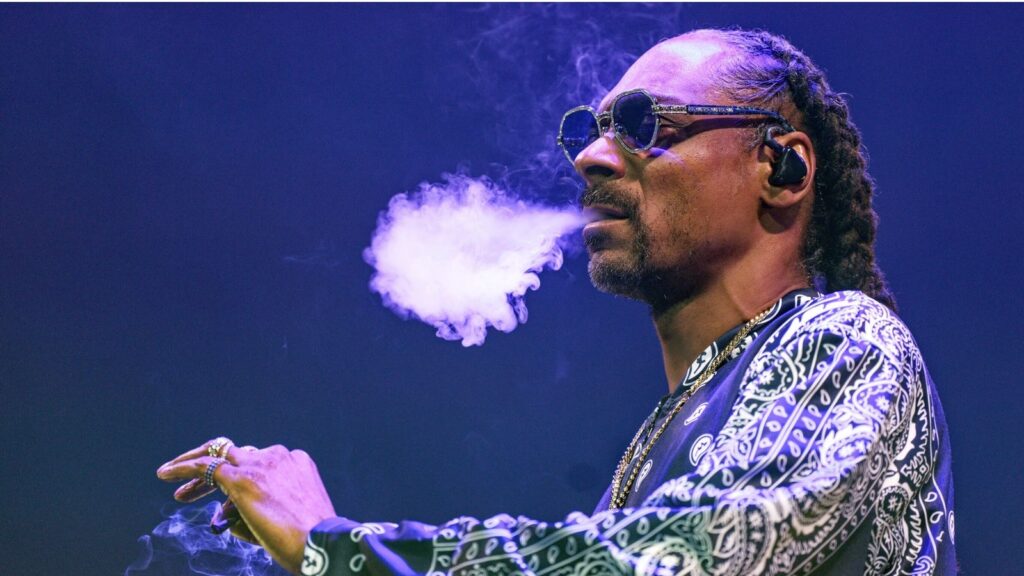 Foto mostra o rapper Snoop Dogg segurando um baseado enquanto expele um trago. Imagem: Henning Kaiser / AP.